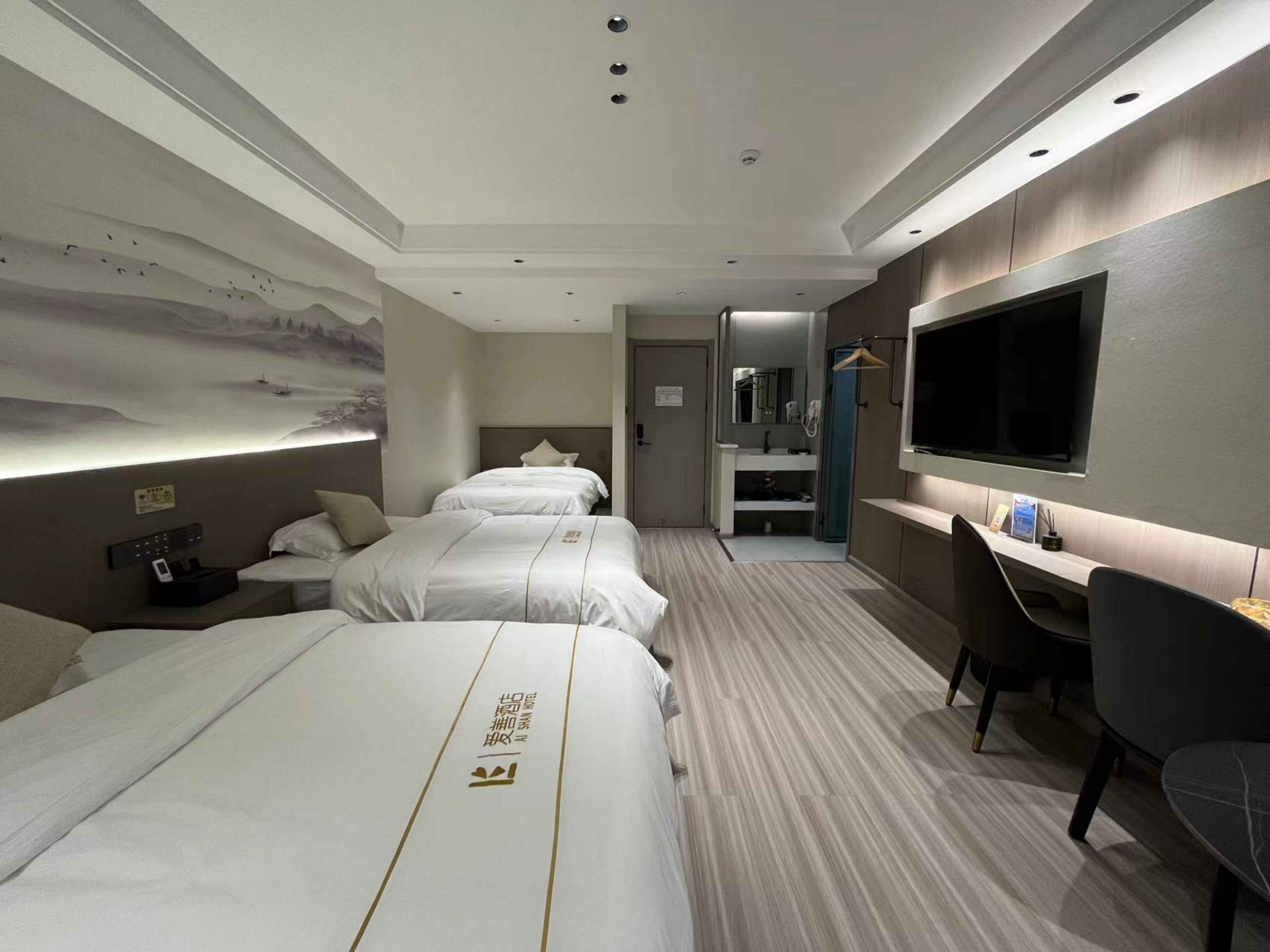 A hotel room with three beds, a desk, a TV, and a bathroom visible through an open door.