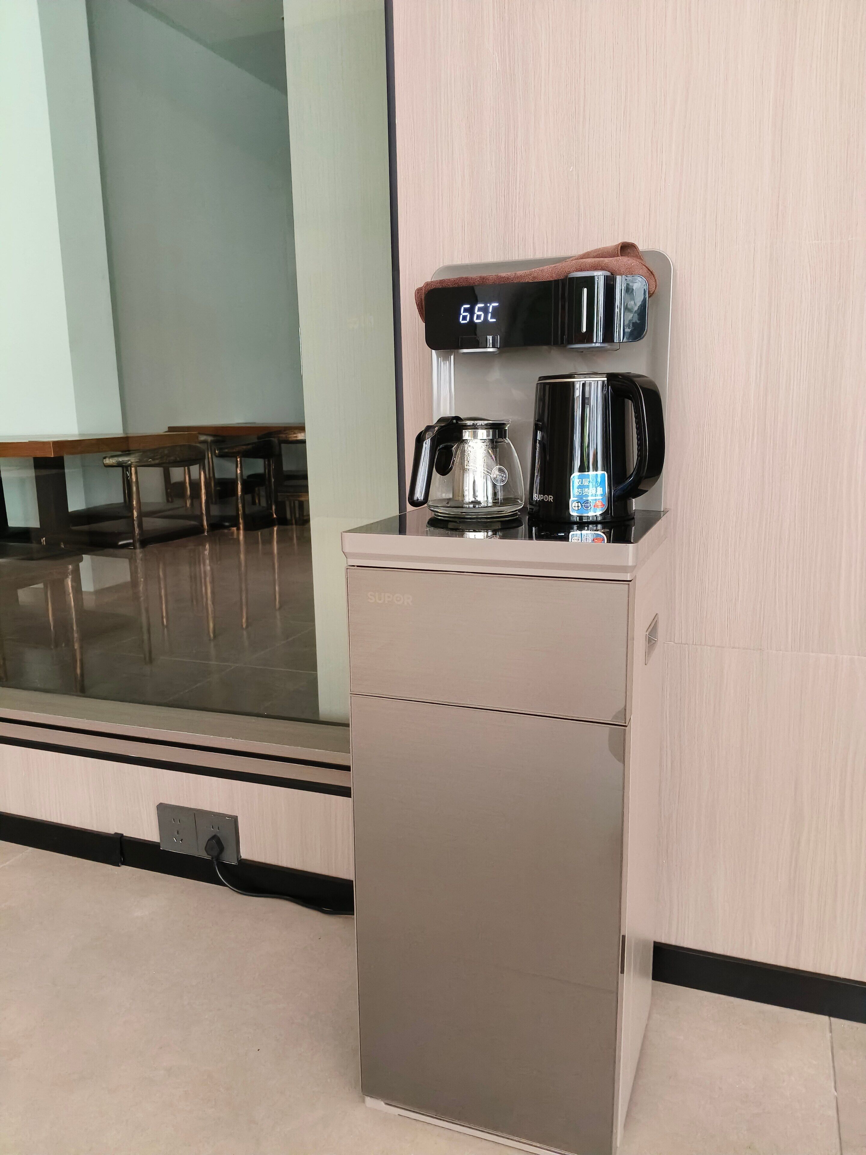 Cafetera compacta con pantalla digital sobre un mueble de pared, jarra de vidrio y recipiente negro para café.