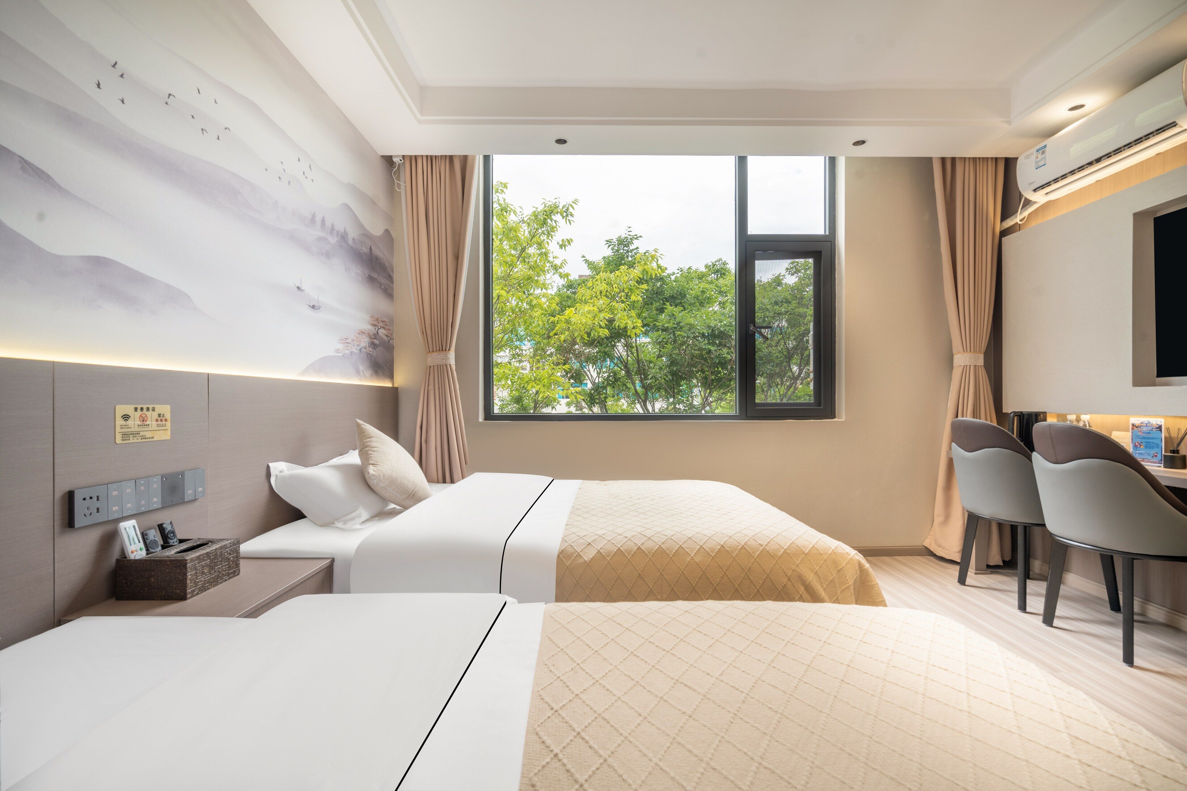 A hotel room with two beds, a desk, chairs, a large window with a view of trees, and a wall-mounted air conditioner.