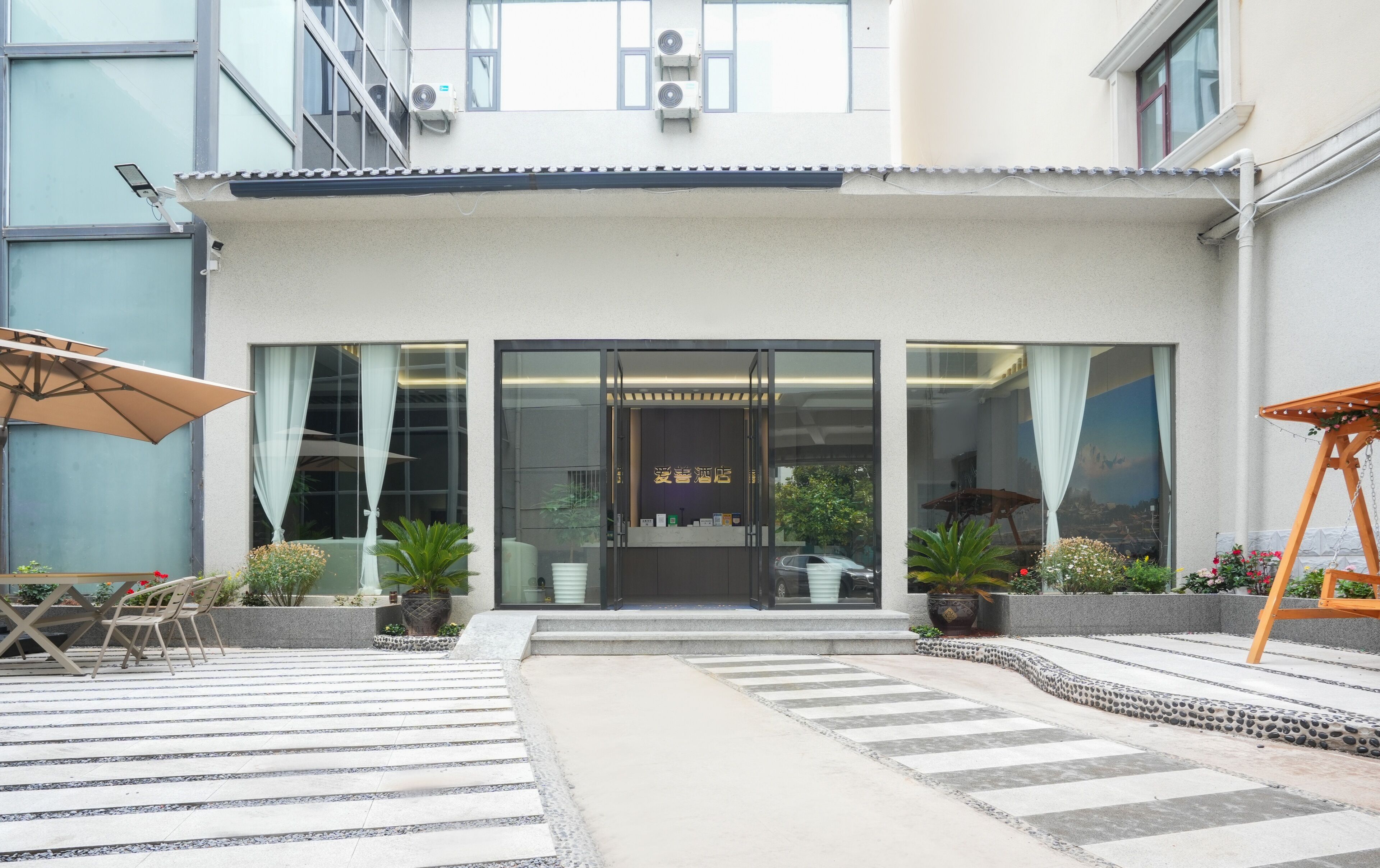 A hotel entrance with a modern design, featuring a reception desk, large windows, and outdoor seating area with umbrellas.