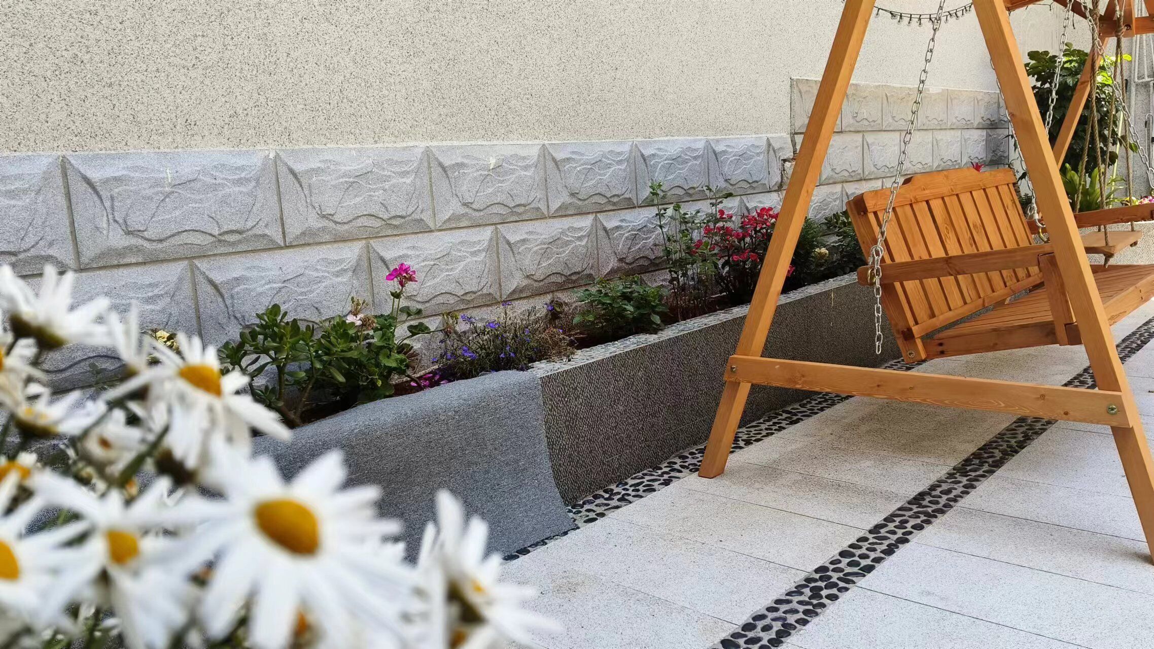 Ein hölzerner Schaukelstuhl in einem Garten mit einer Steinmauer und einem gefliesten Boden.