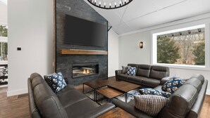 Una sala de estar moderna con sofás de cuero, chimenea, televisor de pantalla plana y un ventanal con vistas a árboles.