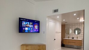 A modern hotel room with a bed, a TV mounted on the wall, a wooden dresser, and a bathroom visible through an open door. - Luxury house with heated pool+Movie theatre room next to sixflags and la cantera (San Antonio)