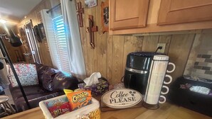 Un área de cocina con armarios de madera, una cafetera, una cesta de aperitivos y un letrero que dice 'Coffee Please'.