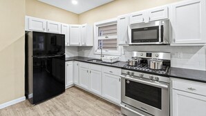 A modern kitchen with white cabinets, a black refrigerator, stainless steel appliances, and a wooden floor. - Duplex|4Min Walk to NYC Train|Gameroom|Hotub+Sauna (Bayonne)