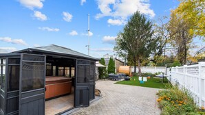 A covered outdoor seating area with a hot tub, a pergola, and a patio with a white fence and greenery. - Duplex|4Min Walk to NYC Train|Gameroom|Hotub+Sauna (Bayonne)
