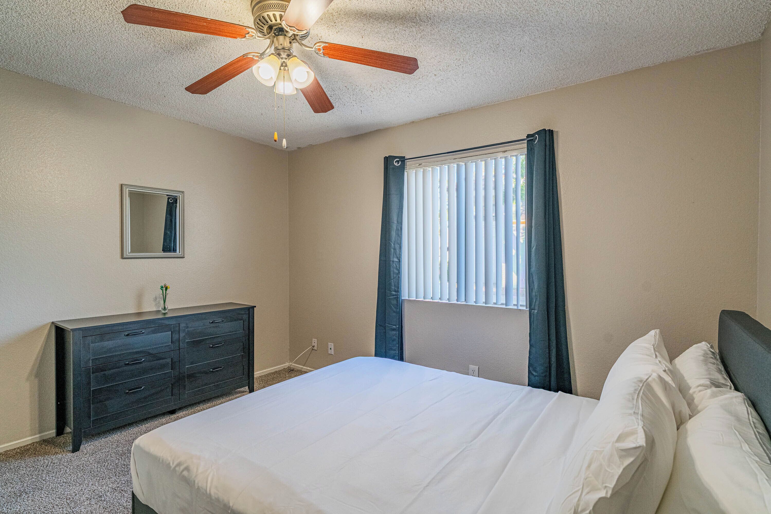 A bedroom with a bed, a dresser, a ceiling fan, a window with curtains, and a wall-mounted mirror.