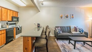 A modern kitchen with wooden cabinets, a dining area with a gray table and chairs, and a gray sofa with a patterned throw.