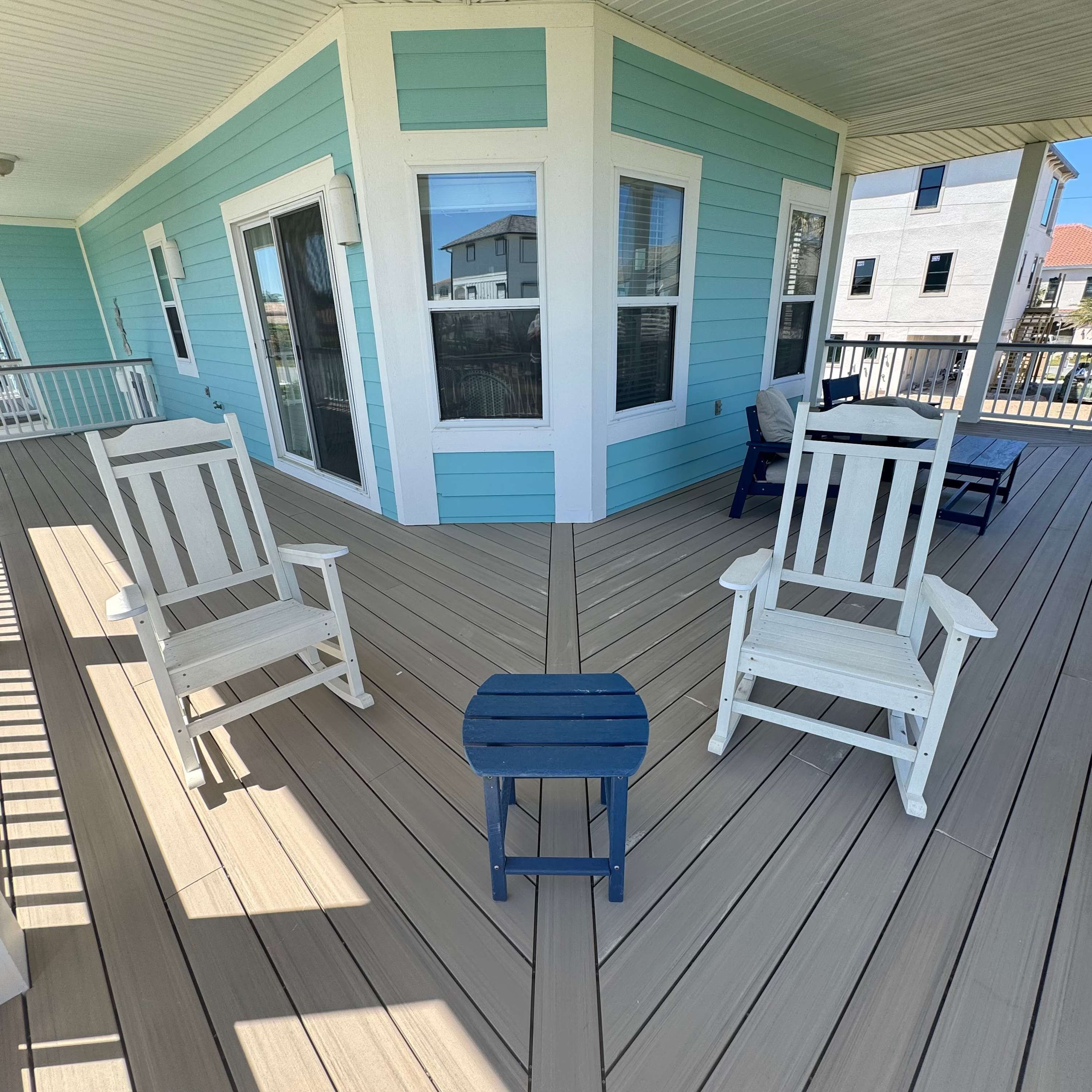 A covered deck with two white Adirondack chairs and a small table, surrounded by teal and white walls with large windows.
