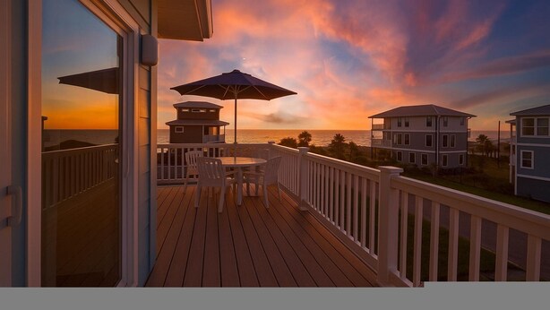 Eine Holzterrasse mit Tisch und Stühlen mit Blick auf einen Sonnenuntergang über dem Meer.