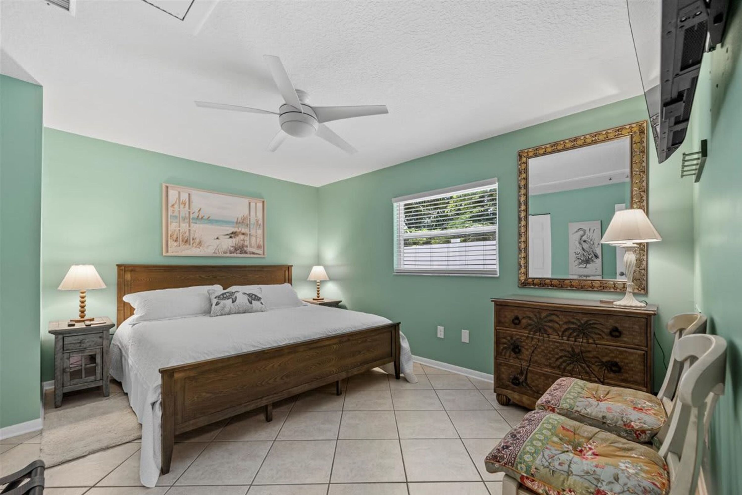 A bedroom with a bed, two bedside tables with lamps, a dresser, a chair, a ceiling fan, and a window with blinds.