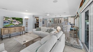 A spacious living room with a large TV, a sofa, and a dining area with a kitchen in the background. - Waterfront Retreat With Pool Tiki Bar Boat Dock (Palm Coast)