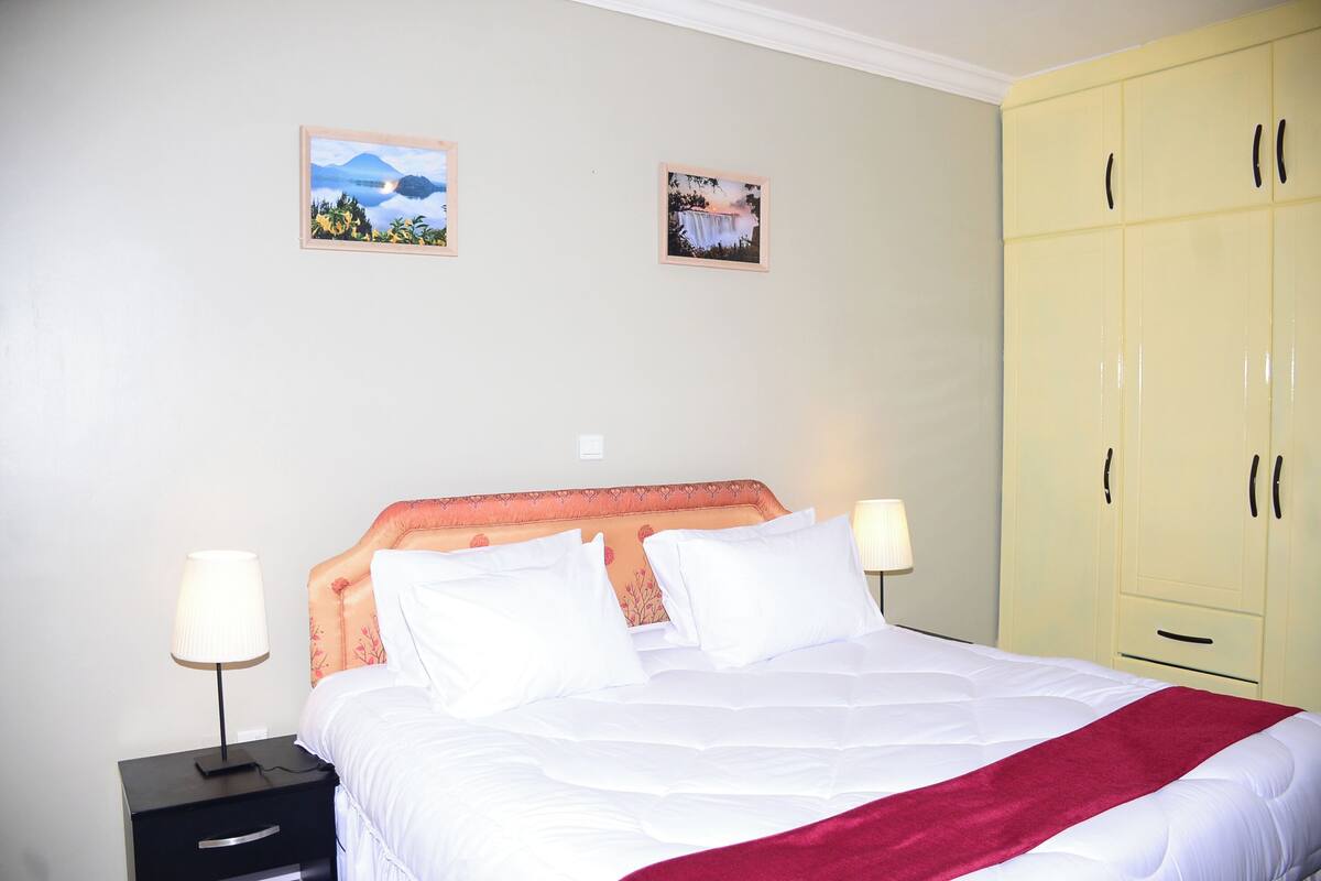 A neatly made bed with white linens and a red throw blanket, a nightstand with a lamp, and a yellow wardrobe.
