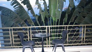 A balcony with a round table and two chairs, surrounded by large banana leaves and a metal railing.