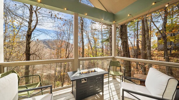 A covered outdoor deck with a fireplace, seating, and a view of the forest. - Cozy Cottage in Yonahlossee Racquet Club (Boone)