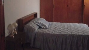 Un dormitorio con un armario de madera, una cama con cubrecama gris, una mesita de noche con lámpara y una cruz en la pared.