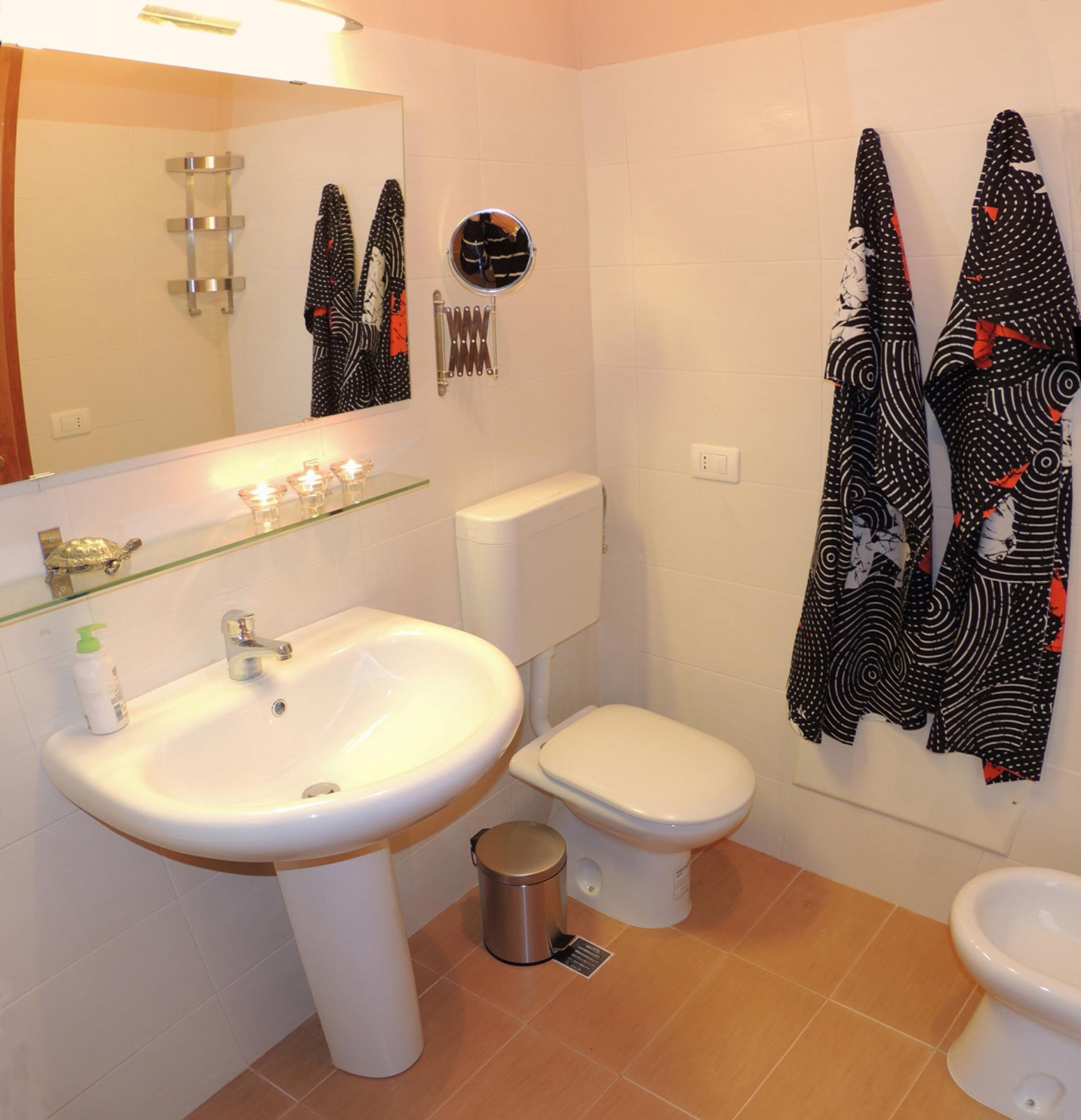 A bathroom with a white sink, toilet, and bidet. There are towels hanging on the wall and a small shelf with toiletries.