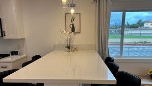 Moderni keittiö, jossa on keskeinen saareke, mustat tuolit, kehystetty taideteos ja ikkuna, josta avautuu näkymä aidalle ja vihreälle kasvillisuudelle.