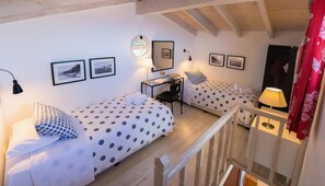 Ein gemütliches Schlafzimmer mit zwei Betten, einem Schreibtisch und einem Sessel. An den Wänden hängen Bilderrahmen und das Fenster bietet einen Ausblick.