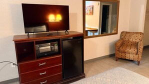 Un espace de vie compact comprenant un meuble de rangement en bois pour les appareils multimédias, un téléviseur à écran plat, un réfrigérateur et un fauteuil à motifs.