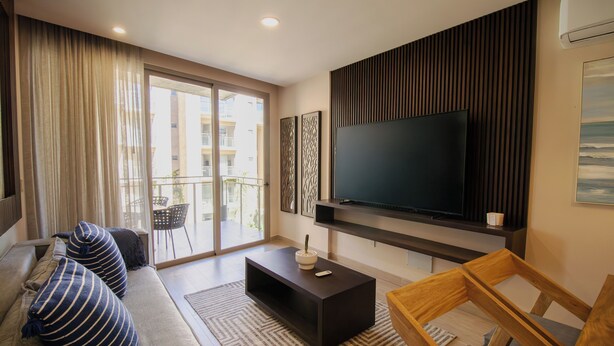 A modern living room with a large flat-screen TV, a sofa with striped cushions, a coffee table, and a wooden chair. - Stay & Relax: Fully Equipped Loft in Los Cabos (Cabo San Lucas)