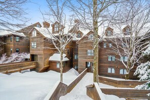A multi-story wooden building with a snow-covered landscape, wooden railings, and a clear sky. - Ski-in queen hotel room with pool, hot tub, and Wi-Fi. (Killington)