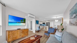 Een moderne woonkamer met een grote televisie, een houten kast, een grijze bank en een eethoek, met een keuken op de achtergrond.