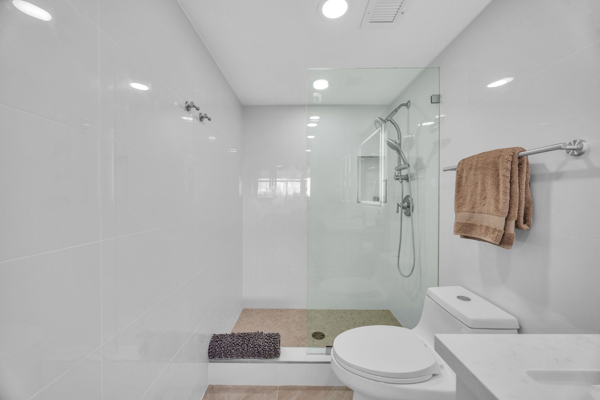 Une salle de bain moderne avec une douche à parois de verre, des toilettes blanches et un tapis de bain beige.