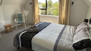 Ein Schlafzimmer mit einem Bett, einem Sessel, einem Standventilator und einem Fenster mit Vorhängen.