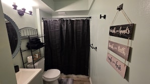 Une petite salle de bain avec un rideau de douche, des toilettes, un lavabo et des étagères contenant des articles de toilette. Des panneaux sont affichés sur le mur pour rappeler de se laver les mains, de se brosser les dents et de se peigner.
