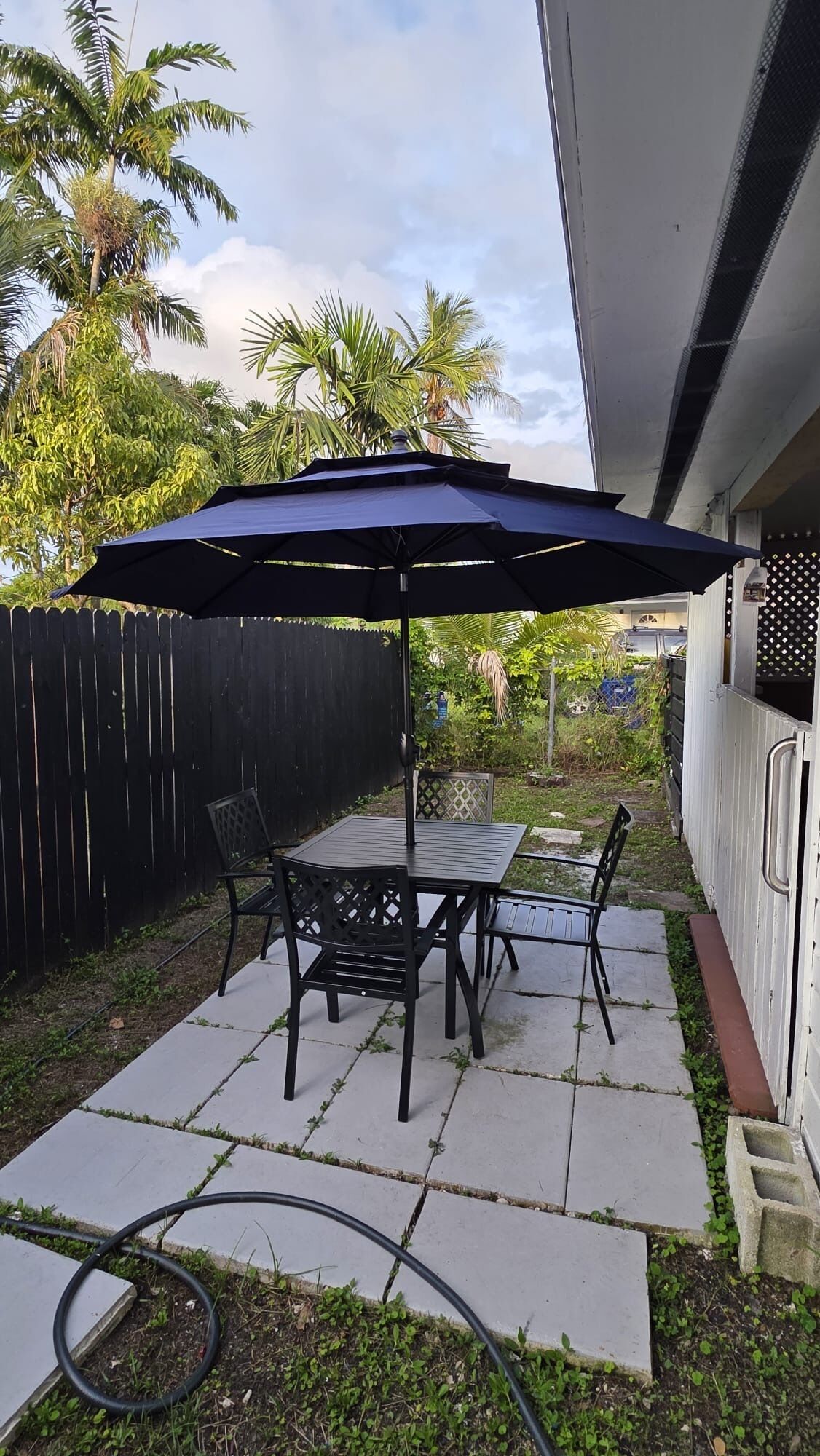 An outdoor patio with a table and chairs under a large umbrella, surrounded by a wooden fence and greenery.