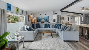 Een moderne woonkamer met een grijze modulaire bank, een salontafel en een groot schilderij van een pier. In de achtergrond is een keuken met een bar en krukken te zien.