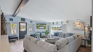 Een ruime woonkamer met een grote grijze bank, een salontafel en een televisie die aan de muur is bevestigd. Er zijn twee stoelen met blauwe kussens, een klein bijzettafeltje met een lamp en een raam met gordijnen.