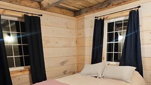Une chambre dans une cabane en bois, avec un lit, deux fenêtres avec des rideaux et un tapis à motifs.