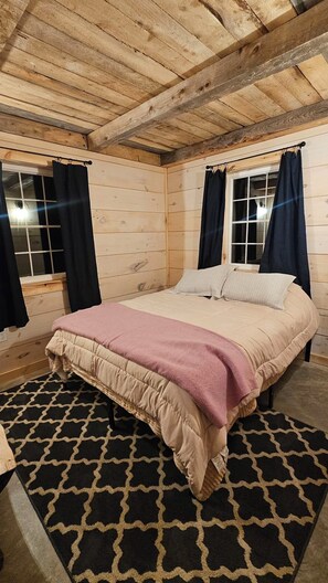 A wooden cabin bedroom with a bed, two windows with curtains, and a patterned rug.