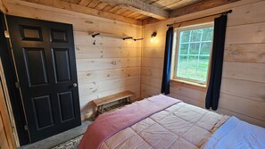 A room with a bed, a bench, a door, and a window with curtains. - Larch - The Tamaracks - Outdoor Adventures Aroostook County (Caribou)