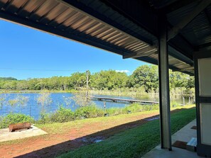 A covered outdoor area with a view of a lake, a wooden dock, and a concrete pathway. - Sunshine Gardens FarmStay (South Mission Beach)
