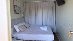 Un lit simple avec un matelas et des oreillers blancs, un téléviseur fixé au mur et un rideau qui recouvre la fenêtre.