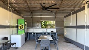 Un espace cuisine extérieur abrité, avec une table et des chaises, un ventilateur de plafond et une vue sur la verdure par une fenêtre.