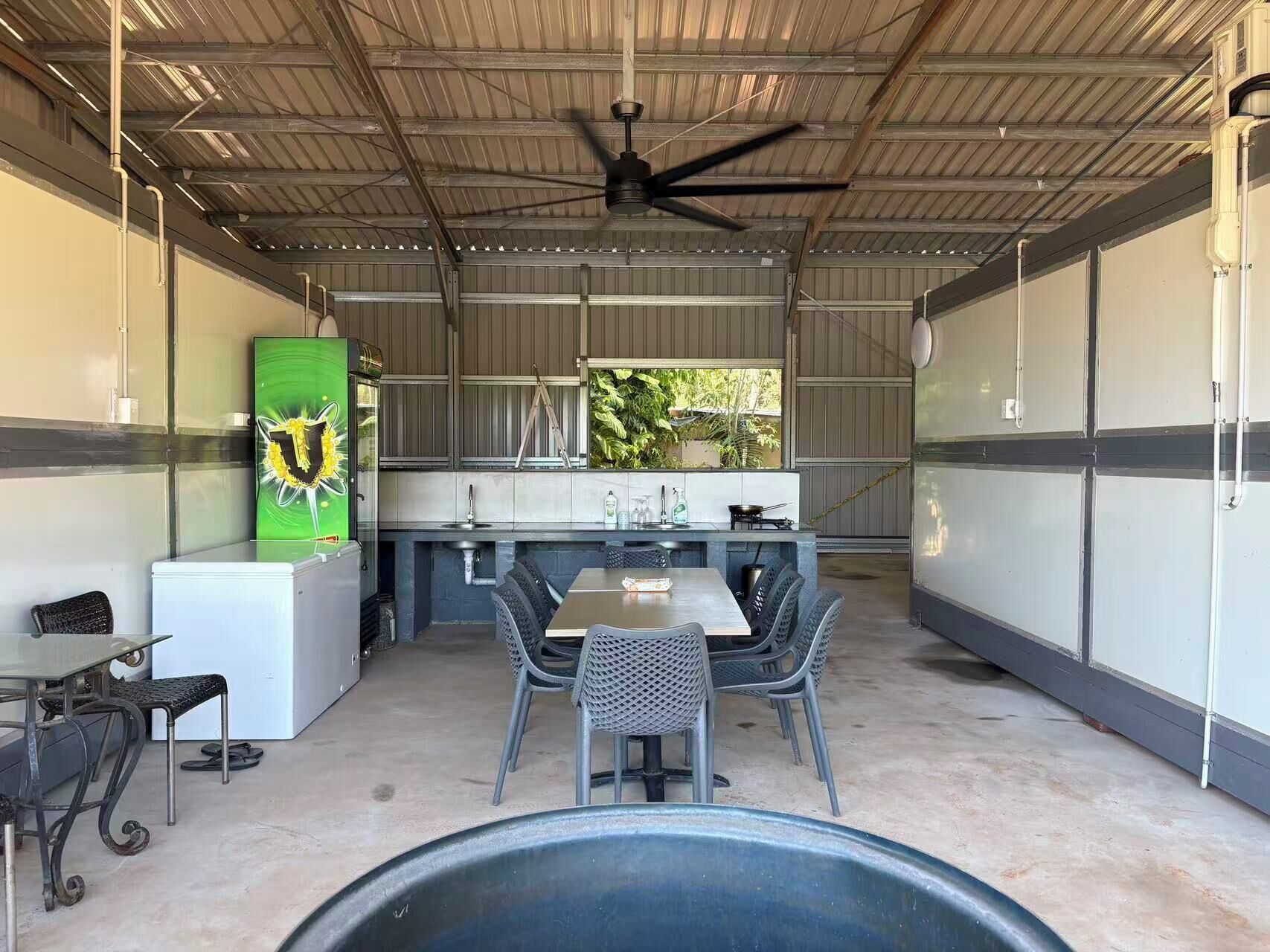 Un espace extérieur abrité, aménagé pour les repas, avec une table et des chaises, un ventilateur de plafond et une vue sur la verdure par une fenêtre.