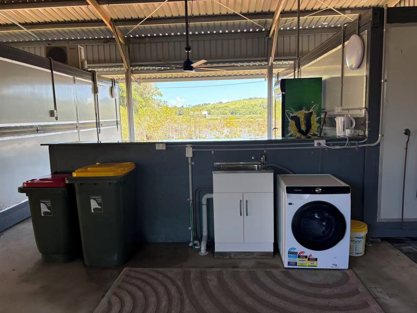 Une cuisine compacte équipée d’un lave-linge, de deux poubelles, d’un petit meuble et d’une fenêtre donnant sur un espace verdoyant.