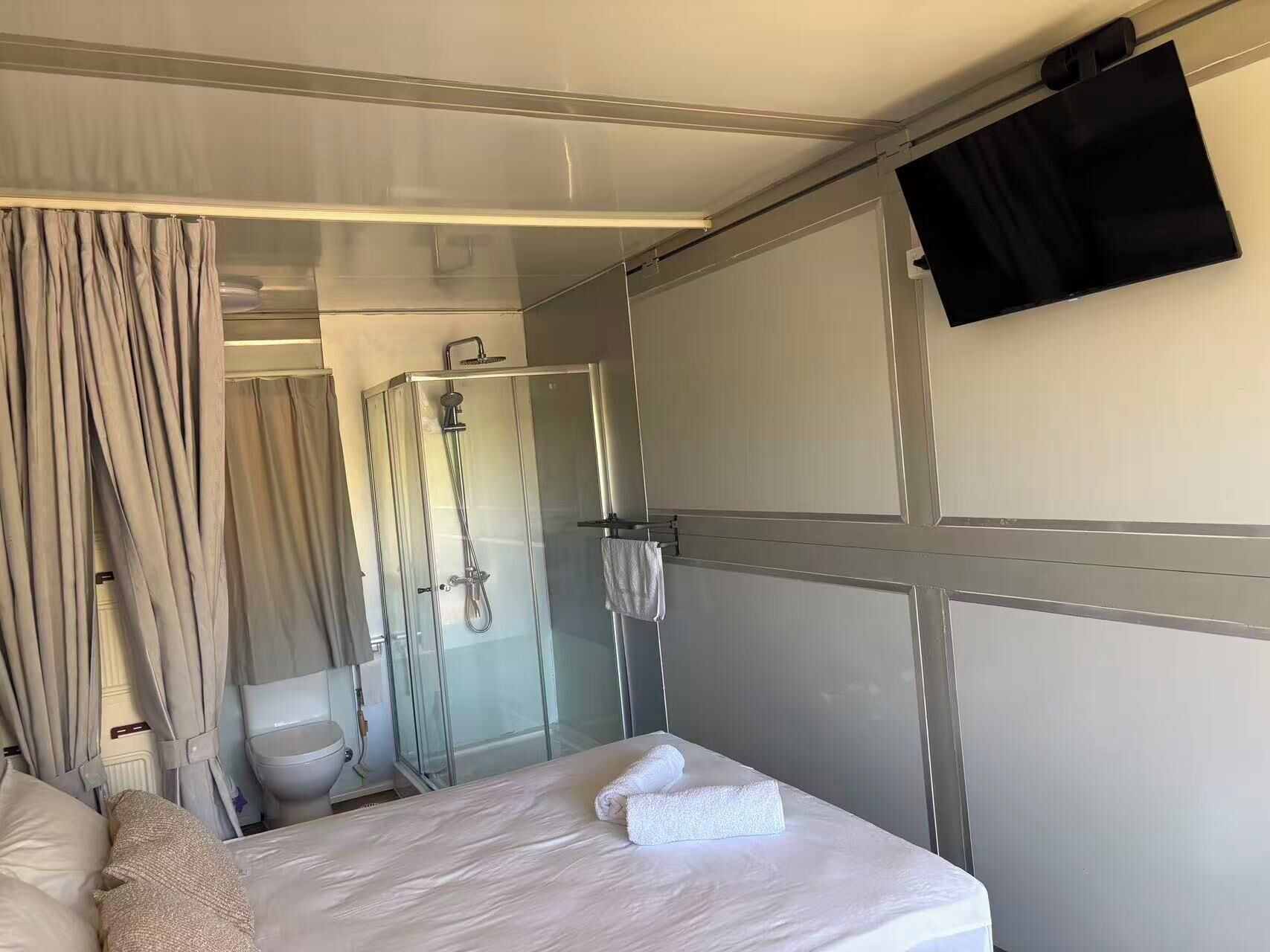 A hotel room with a bed, a shower, a toilet, and a TV mounted on the wall.