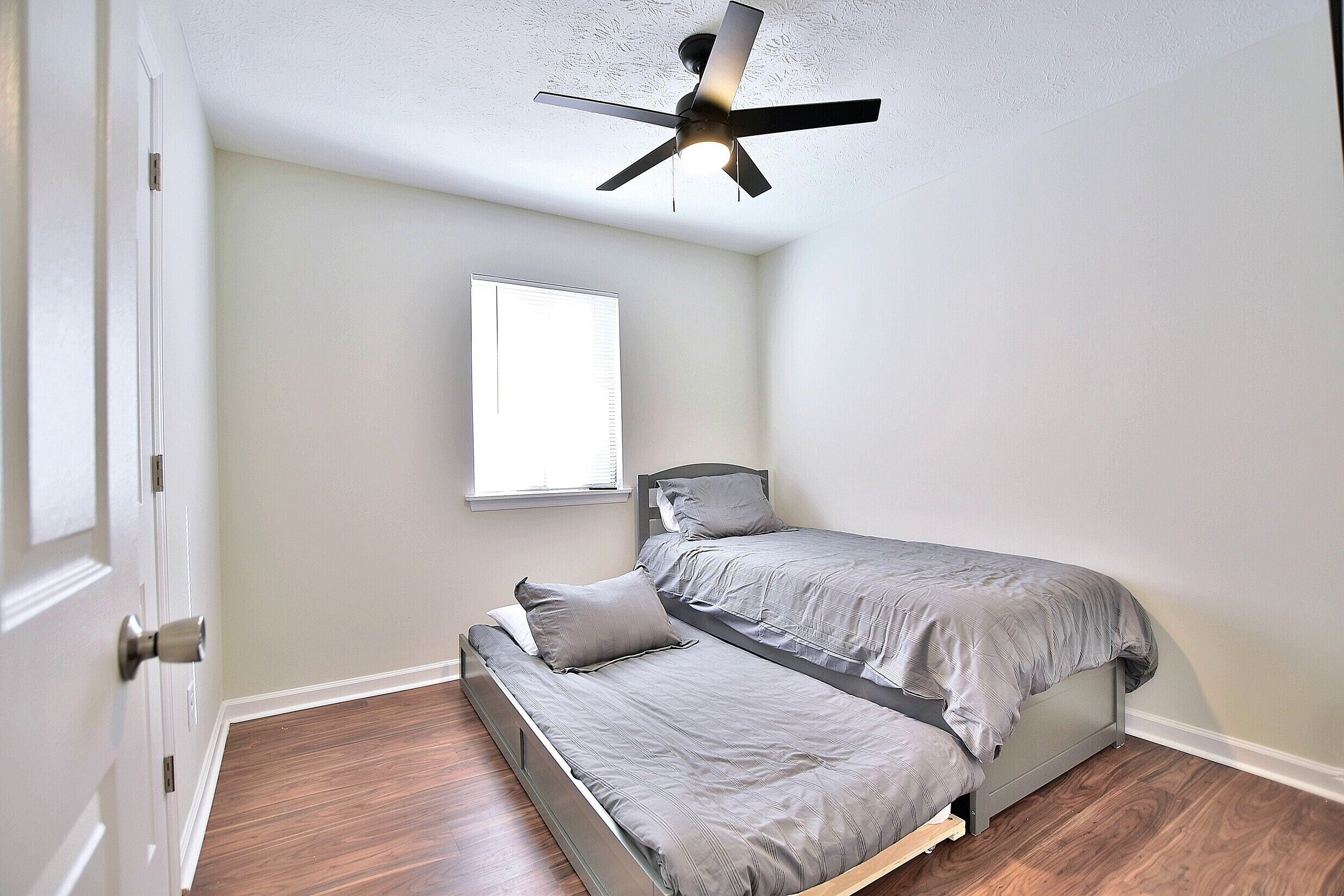 Une chambre avec un lit simple, un ventilateur de plafond, une fenêtre et une porte.