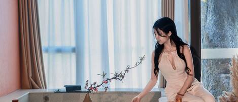 A woman sitting on a stone bench in a modern room with a large window, a vase with branches, and a coffee table with books and a cup.