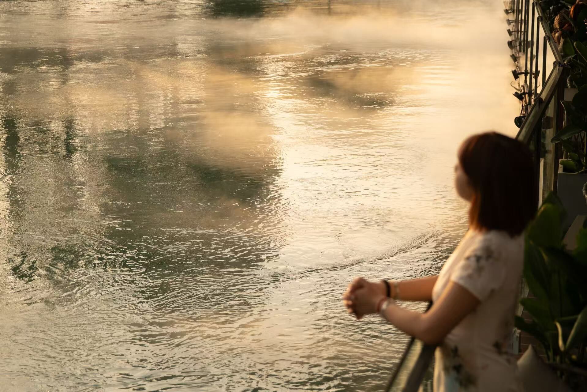 A person standing by a calm body of water, possibly a river or lake, with a railing on the right side.