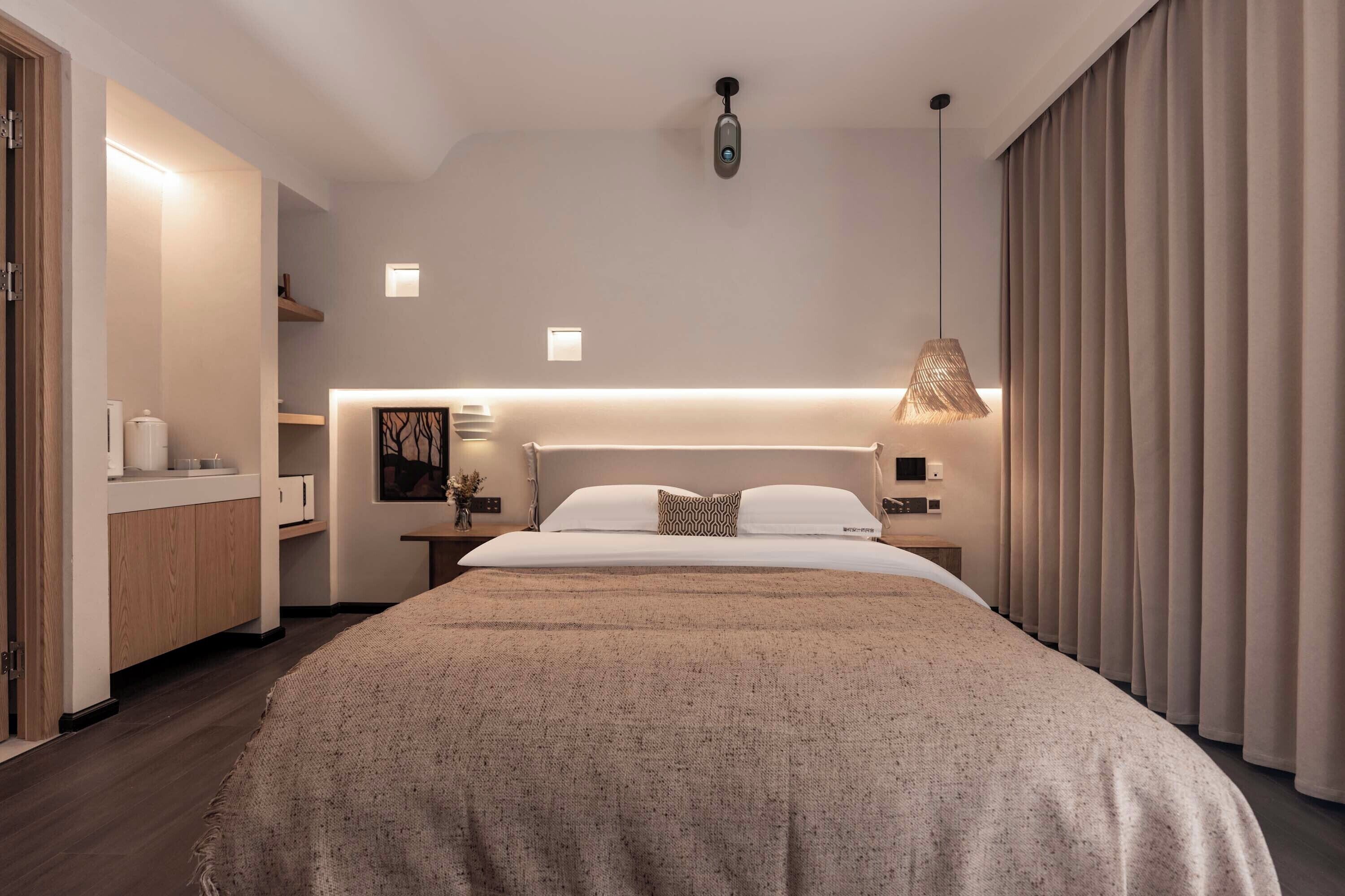 A modern hotel room with a large bed, bedside tables, a nightstand, a hanging light fixture, and a view of the outside through the window.