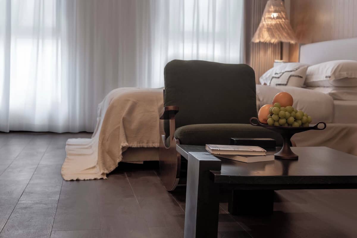 A modern hotel room with a dark armchair, a small table with a fruit bowl, and a bed with a white cover.