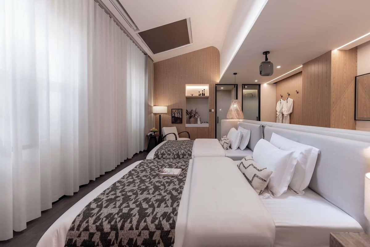 A modern hotel room with two beds, a sitting area, and a bathroom with a mirror and towel rack.