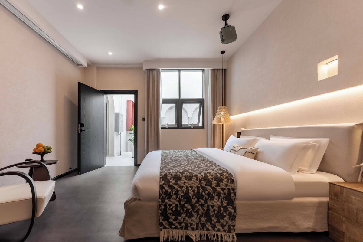 A hotel room with a large bed, bedside table, lamp, and a view of the outside.
