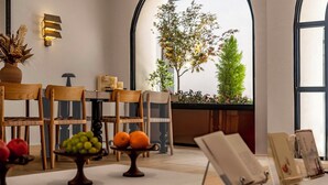 Uma área de refeições com cadeiras de madeira, uma mesa com frutas, um vaso e vista para árvores através de janelas amplas.